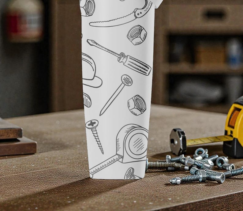 May include: A white tumbler featuring a repeating pattern of hand-drawn tools, including a saw, screwdriver, and tape measure. The tumbler is set on a wooden surface with additional tools and hardware in the background.