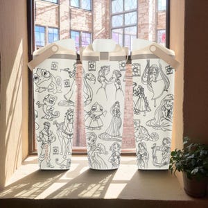 Peut inclure: Trois bouteilles d'eau blanches avec une poignée et un couvercle beiges, chacune ornée d'un dessin au trait noir de princesses et de personnages Disney. Les bouteilles sont rectangulaires et posées sur un rebord de fenêtre. Le mot "owala" est visible sur une bouteille.