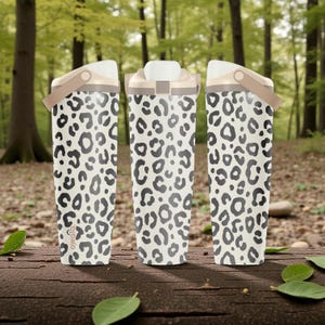 Puede incluir: Tres botellas de agua Owala con base crema y estampado de leopardo negro. Cada botella tiene una tapa blanca, un asa beige y una apertura con botón. Las botellas se exhiben sobre una superficie de madera con un fondo de bosque.