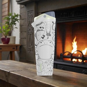 May include: White water bottle with a black and white cartoon design featuring a frog, hot dog, taco, and other food items. The bottle has a gray lid and a white handle. The bottle is sitting on a wooden surface.
