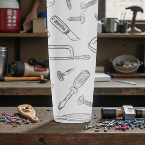 May include: A white tumbler featuring a repeating pattern of hand-drawn tools, including screwdrivers, saws, and nails. The tumbler is cylindrical and stands upright on a wooden surface, with various tools and hardware scattered around it.