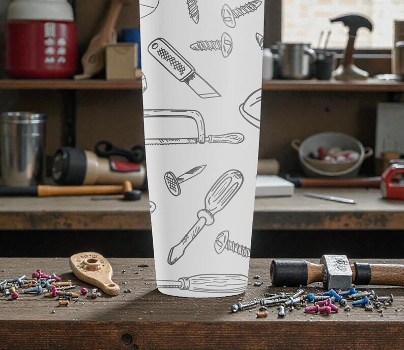 May include: A white tumbler featuring a repeating pattern of hand-drawn tools, including screwdrivers, saws, and nails. The tumbler is cylindrical and stands upright on a wooden surface, with various tools and hardware scattered around it.