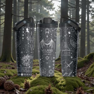 Puede incluir: Tres vasos de viaje negros con un diseño gráfico blanco. Cada vaso presenta una ilustración diferente, incluyendo un ojo de dragón, una criatura alada y un castillo. El texto "VELARIS city of starlight" está impreso en los vasos.