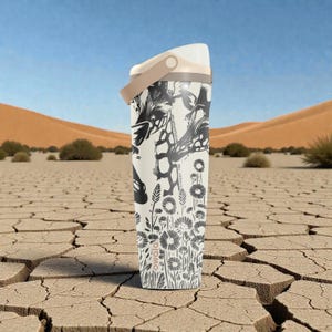以下が含まれることがあります： 白と黒のOwalaウォーターボトル。花柄とキリンのデザイン。ボトルにはベージュのハンドルと蓋が付いています。背景には、ひびの入った土と青い空のある砂漠の風景が写っています。