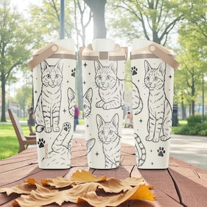 Peut inclure: Trois bouteilles d'eau isolées blanches avec des couvercles et des poignées beiges. Chaque bouteille présente une illustration de chat noir et blanc, des empreintes de pattes et des étoiles. Le mot "owala" est imprimé sur une bouteille. Les bouteilles sont sur une table en bois avec des feuilles d'automne.