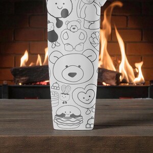 May include: A white tumbler featuring a repeating pattern of cartoon bears and other cute characters. The design is in black outline on a white background. The tumbler is set against a backdrop of a fireplace with a burning fire.
