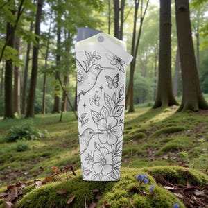 Pu&ograve; includere: Borraccia bianca con un design di colibr&igrave; e fiori in bianco e nero. La borraccia ha un coperchio grigio e un manico bianco. Lo sfondo &egrave; una scena di foresta con muschio verde e alberi.