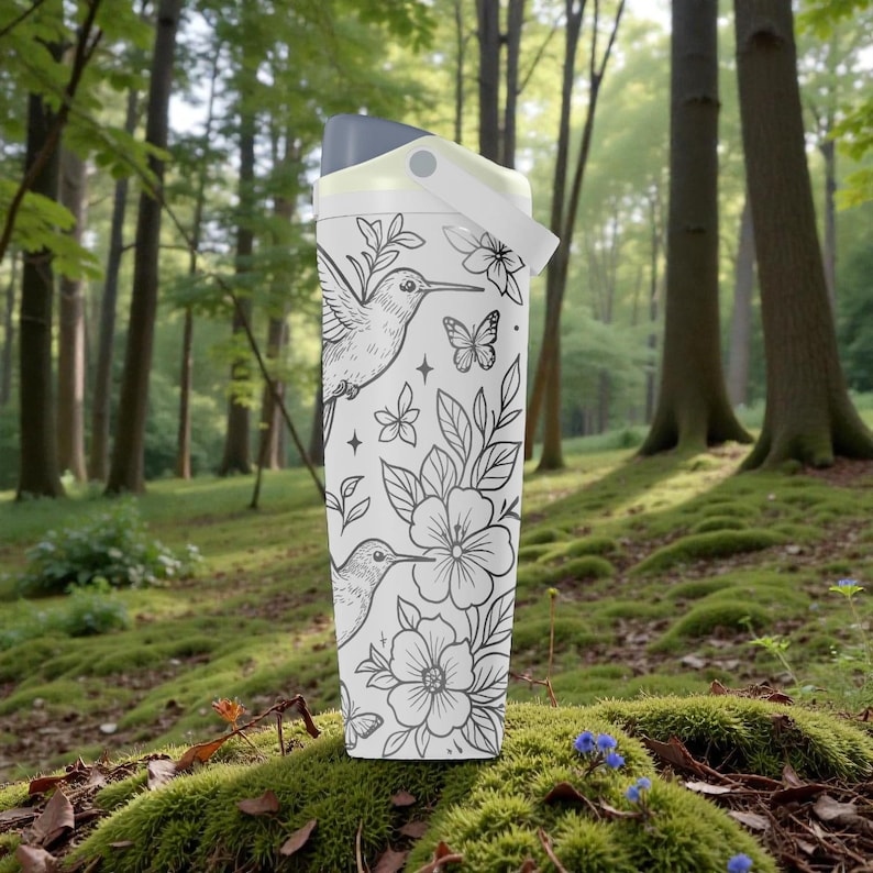Pu&ograve; includere: Borraccia bianca con un design di colibr&igrave; e fiori in bianco e nero. La borraccia ha un coperchio grigio e un manico bianco. Lo sfondo &egrave; una scena di foresta con muschio verde e alberi.
