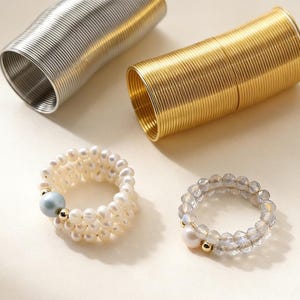 以下が含まれることがあります： 2 つのリングと 2 つのコイル状金属チューブを含む、さまざまなジュエリー。1 つのリングは、小さな白い真珠と水色のビーズでできています。もう 1 つのリングは、透明なファセットビーズと白い真珠でできています。金属チューブは金と銀です。