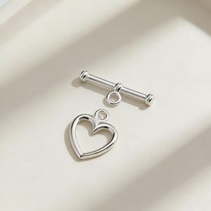 以下が含まれることがあります： ハート型のペンダントと丸みを帯びた端を持つバーを備えた、銀色のトグルクラスプセット。 クラスプはジュエリー作りのために設計されており、明るい色の表面に表示されています。