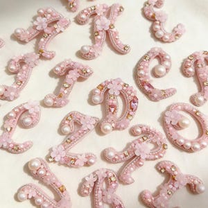 May include: A selection of pink, beaded letters, embellished with pearls, tiny flowers, and gold details. The letters are displayed on a white surface, highlighting a delicate, handcrafted appearance. These decorative letters are perfect for personalised crafts.