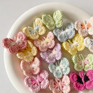 Puede incluir: Colección de adornos de mariposas de ganchillo de colores pastel, rosa, amarillo, verde y azul. Las mariposas están dispuestas sobre una superficie redonda blanca, mostrando sus delicados detalles hechos a mano.