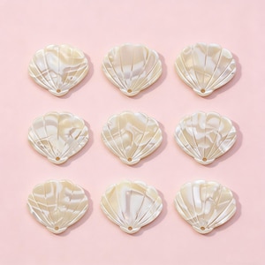 Pode incluir: Nove contas em forma de concha, cor creme, com acabamento perolado, dispostas sobre um fundo rosa. Cada conta tem um design em forma de leque com um pequeno orifício na parte inferior. As contas são provavelmente para fazer joias ou projetos de artesanato.