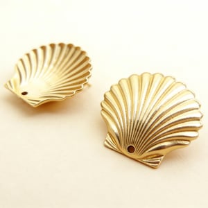 May include: Two gold-colored seashell stud earrings. Each earring is shaped like a scallop shell with detailed ridges radiating from the center. The earrings have a small hole near the base and a post for securing them.