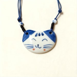 Puede incluir: Un collar con colgante de gato de cerámica blanca con detalles azules. La cara del gato tiene una expresión sonriente, bigotes azules y mejillas rosadas. El collar tiene un cordón azul con una sección negra y cuentas blancas.