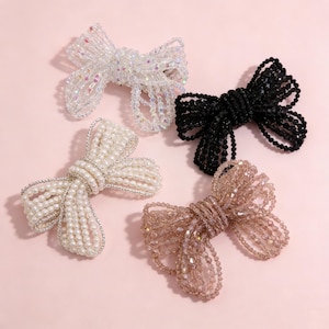 Puede incluir: Cuatro pinzas para el pelo con lazo de cuentas en blanco, negro, perla y rosa rubor. Los lazos están hechos de pequeñas cuentas y están dispuestos en forma de lazo. Las pinzas están sobre un fondo rosa claro.