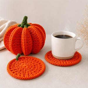 May include: A crocheted orange pumpkin with a green stem, a flat circular orange crocheted coaster with a small green stem, and a white ceramic mug filled with dark coffee resting on a second matching orange crocheted coaster.