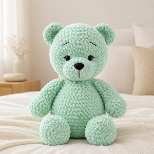 Op de afbeelding: Een mintgroene gehaakte teddybeer gemaakt van dik, pluche garen. De beer heeft een ronde kop met kleine oortjes, een zwarte geborduurde neus, een verticale mondlijn en donkere, ronde ogen met daarboven gestikte wenkbrauwen.