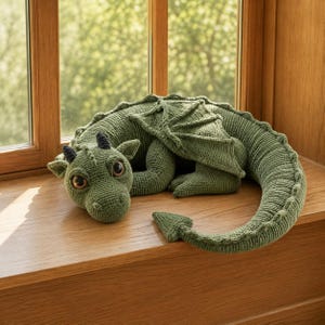 May include: A green crocheted dragon plush toy featuring large brown eyes, two small black horns, textured wings, and a long tail with a pointed tip. The creature is crafted with a detailed knit pattern and rests in a curled position.