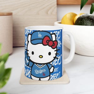 May include: White ceramic mug featuring a cartoon character in a blue baseball uniform with the text "Dodgers". The character wears a blue cap and a red bow. The mug has a blue background with the word "Dodgers" repeated.