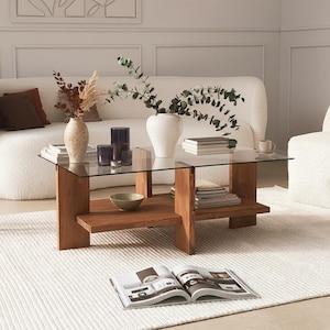 Peut inclure: Un salon avec une table basse en verre et bois, un canapé blanc et un tapis blanc. La table présente des vases avec des plantes et des livres. Un petit tabouret en bois et un fauteuil blanc sont également présents. L'ambiance est neutre.