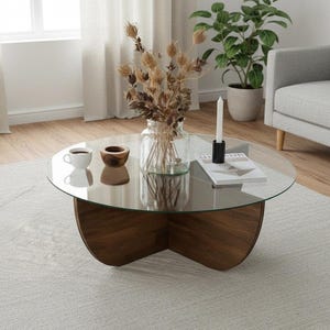 May include: A circular coffee table with a clear glass top and a dark brown wooden base. The table displays a vase of dried flowers, a candle, a small cup, and a wooden bowl. The table rests on a light grey rug in a living room.