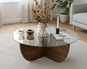 Puede incluir: Una mesa de centro redonda con una tapa de cristal transparente y una base de madera marrón oscuro. La mesa tiene un jarrón con flores secas, una vela, una taza pequeña y un cuenco de madera. La mesa está sobre una alfombra gris claro en un salón.