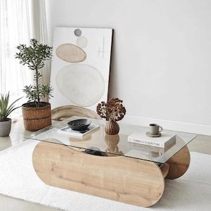 May include: A contemporary coffee table featuring a glass top and a light wood base. The base has a distinctive, curved shape. The table is in a bright room with decorative plants and artwork. A book and a cup are on the table.