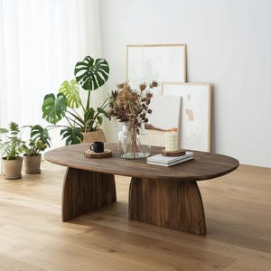 May include: A dark brown, oval-shaped wooden coffee table with a natural finish. The table has a unique base design and is surrounded by plants, artwork, and decorative items. The table is approximately 60 inches long.
