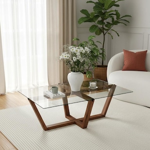 Puede incluir: Una mesa de centro moderna con tapa de cristal y base de madera oscura. La mesa tiene una superficie rectangular de cristal sostenida por un marco de madera geométrico. Un jarrón blanco con flores, una taza y una vela están sobre la mesa.
