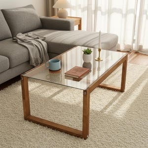 Op de afbeelding: Een moderne salontafel met een glazen blad en een houten frame. Een blauwe mok, een kleine plant in een pot, een boek en een kandelaar met een brandende kaars staan op de tafel. De tafel staat op een lichtgekleurd tapijt in een woonkamer.