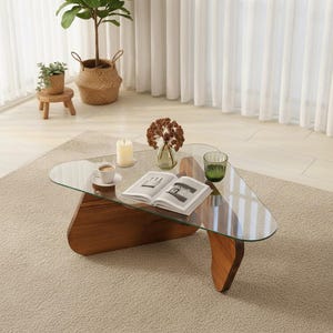 Peut inclure: Une table basse triangulaire moderne avec un plateau en verre transparent et une base en bois. La table est posée sur un tapis beige et comprend une tasse de café, une bougie, un vase avec des fleurs séchées et un livre. La table mesure environ 100 cm de long.