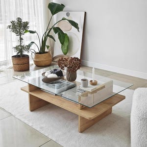 Peut inclure: Table basse moderne avec plateau en verre transparent et base en bois brun clair. De forme rectangulaire, elle est posée sur un tapis blanc. Décorée avec des plantes, des livres et un vase.