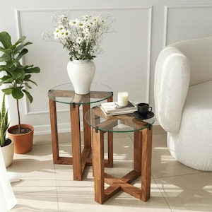May include: Two wooden side tables with clear glass tops. One table has a white vase with white flowers. The other table has a candle, a book, and a black teacup. The tables are made of brown wood.