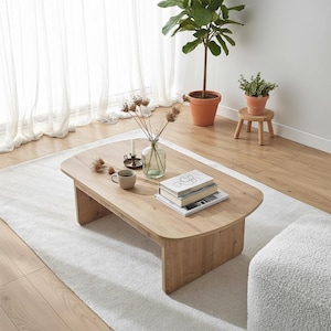 Könnte beinhalten: Ein helles Holz-Couchtisch mit abgerundeten Kanten, auf dem Bücher, eine Tasse und eine Vase mit getrockneten Blumen stehen. Der Tisch steht auf einem weißen Teppich in einem hellen Raum. Eine Topfpflanze und ein kleiner Holzschemel sind im Hintergrund.