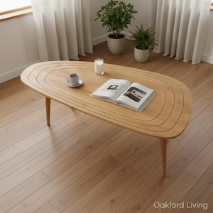 Peut inclure: Une table basse en bois clair de forme arrondie unique, avec une série de rainures incurvées. La table a quatre pieds fins et un magazine, une tasse et un verre sur le dessus. Le texte "Oakford Living" est visible.