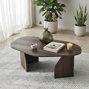 May include: A dark brown oval coffee table with a modern design. The table has a wooden surface and a unique base. A vase, book, candle, and cup sit on the table. The table is surrounded by plants.