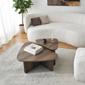 May include: Two dark brown, triangular coffee tables with a wood grain finish. One table is partially under the other. A stack of books sits on the larger table. A white sofa and rug are in the background.