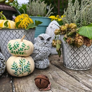 May include: A rustic outdoor scene featuring decorative pumpkins, a concrete owl statue, and potted plants. Two white pumpkins are stacked, adorned with green leaf designs. A small, rusty bell with a red and white string sits nearby.