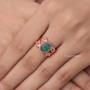 Puede incluir: Un anillo de cobre con una piedra preciosa verde de forma ovalada engastada en un bisel de metal trenzado. La banda está decorada con intrincados adornos metálicos y pequeños acentos esféricos a ambos lados de la piedra. El texto CopperiaCo es visible.
