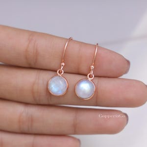 May include: A pair of drop earrings featuring round, iridescent moonstone cabochons set in rose gold-toned metal. Each earring has a simple hook wire attachment. The text CopperiaCo is visible in the lower right corner of the image.