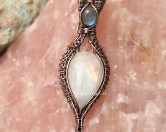 Peut inclure: Un pendentif artisanal avec une pierre précieuse irisée en forme de poire et une petite pierre bleue ovale, serti dans un tissage de fil de cuivre. Le pendentif est présenté sur une surface de pierre rose. Le mot "Copper&Co" est visible.