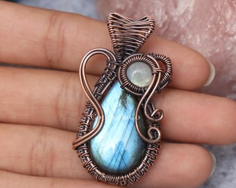 Peut inclure: Un pendentif en fil de cuivre torsadé orné d'une pierre de labradorite en forme de goutte aux reflets bleus et d'une petite pierre de lune ronde. L'ensemble présente des volutes et des enroulements métalliques complexes autour des pierres.