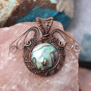 Könnte beinhalten: Ein Anhänger mit einem runden, irisierenden Abalone-Muschel-Cabochon, der in eine komplexe Umwicklung aus dunklem Kupferdraht eingefasst ist. Das Drahtdesign bildet symmetrische, flügelartige Verzierungen und eine zentrale Öse. Der Text CopperiaCo ist sichtbar.