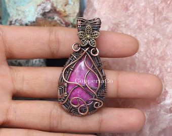 Peut inclure: Un pendentif artisanal avec une pierre précieuse rose vif en forme de goutte d'eau. La pierre est enfermée dans un tissage de fil de cuivre avec des volutes décoratives et un accent floral. Le pendentif porte le texte "Copperiaco".