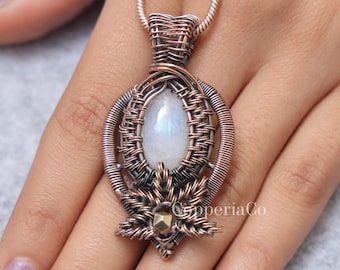 Peut inclure: Un collier pendentif artisanal en fil de cuivre avec une pierre de lune irisée en forme de poire. Le pendentif présente un design tissé complexe avec une base florale et un petit accent doré. Le collier est présenté sur une surface gris clair.