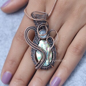 May include: A handcrafted copper wire pendant necklace. The pendant features an oval-shaped iridescent abalone shell, a small round stone, and intricate wire wrapping. The necklace is displayed on a hand.