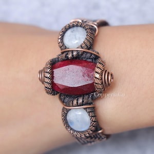 May include: A handcrafted copper bracelet featuring a large, faceted, deep red gemstone at the center, flanked by two smaller, round, pale blue gemstones. The bracelet has a woven design with intricate wirework.
