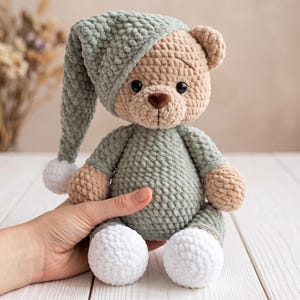 May include: A crocheted teddy bear made of soft, textured yarn in shades of beige, sage green, and white. The bear wears a matching sage green nightcap with a white pom-pom and a sage green onesie, featuring dark brown eyes and a brown nose.