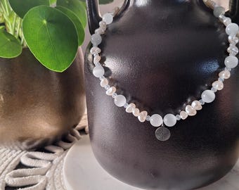 May include: A delicate necklace featuring a mix of white and pale blue beads, and small, irregular pearls. The necklace is draped around a dark, matte black vase. A small, silver-colored charm hangs from the center of the necklace.